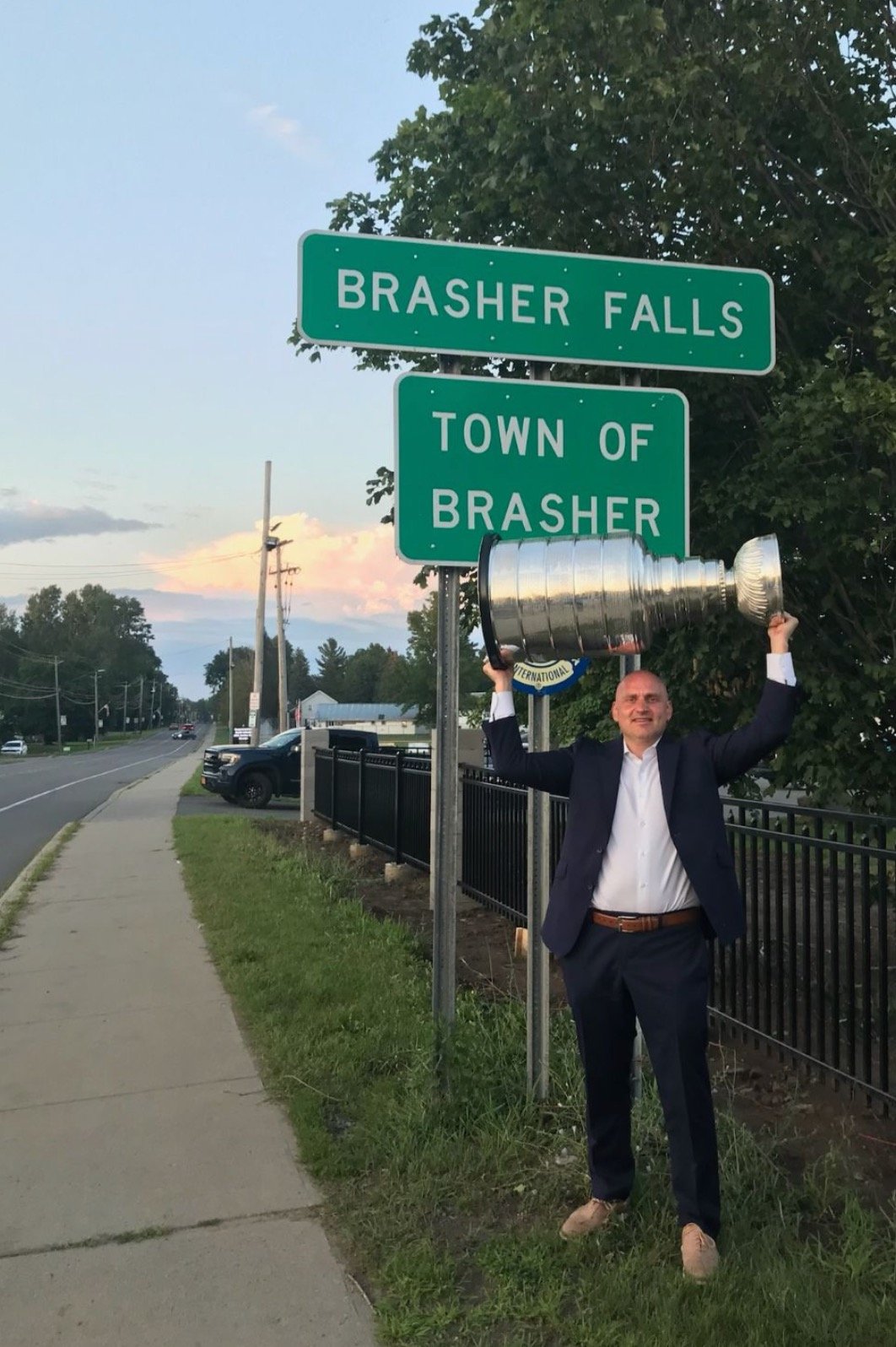 A man in a suit holding a large replica hockey trophy with both hands while standing next to street signs that read 'BRASHER FALLS' and 'TOWN OF BRASHER.' The scene is outdoors along a sidewalk with trees and parked cars in the background during sunset.