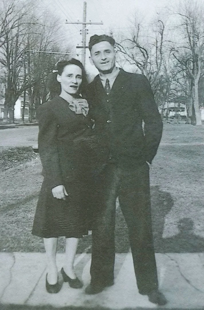 A black and white photo of a young couple standing outdoors on a sidewalk, with leafless trees and houses in the background. The woman is wearing a dark dress with a large bow at the neckline and heels, while the man is dressed in a dark suit with a shirt and tie. They are standing close, smiling at the camera.
