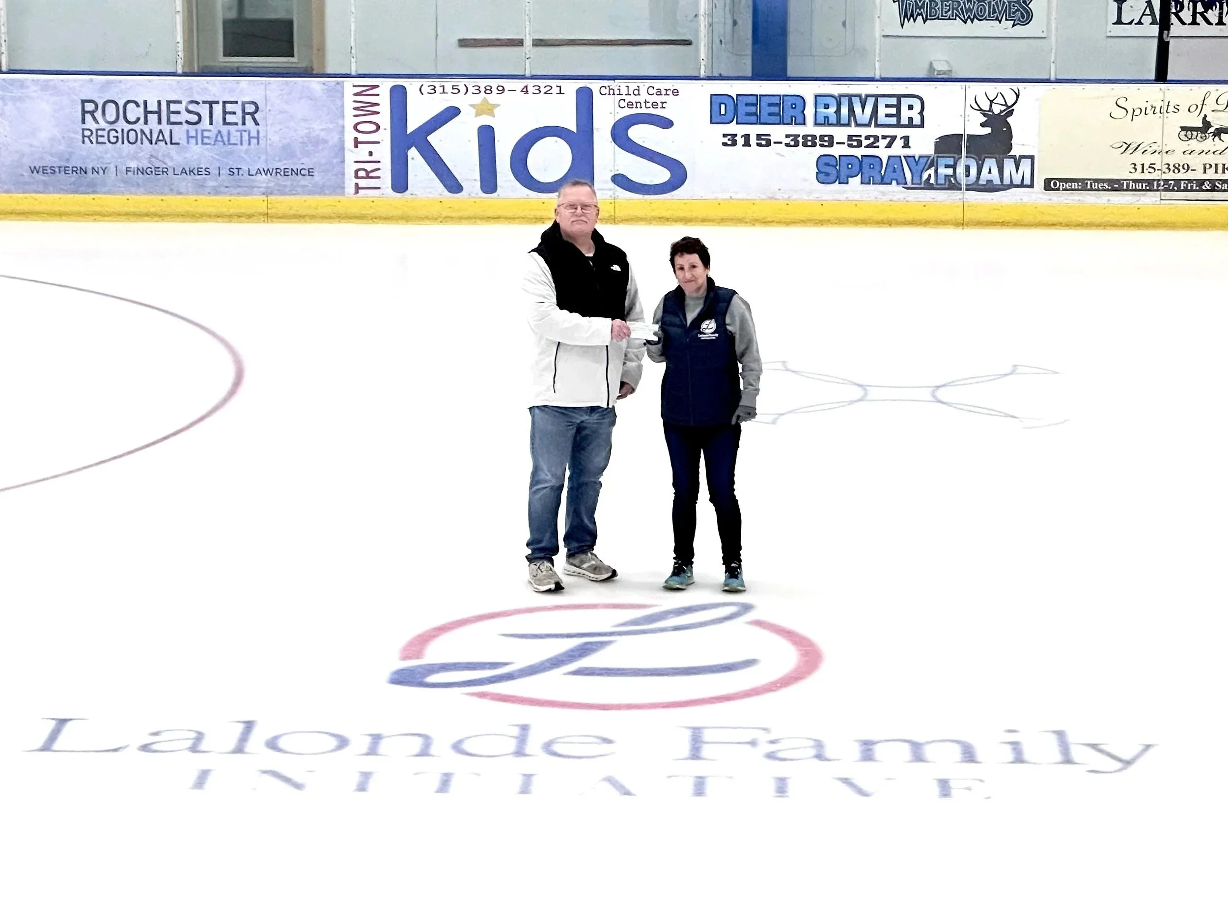 Two people standing on an ice hockey rink, one man and one woman, near the center logo, with advertisements and signs visible on the boards behind them.