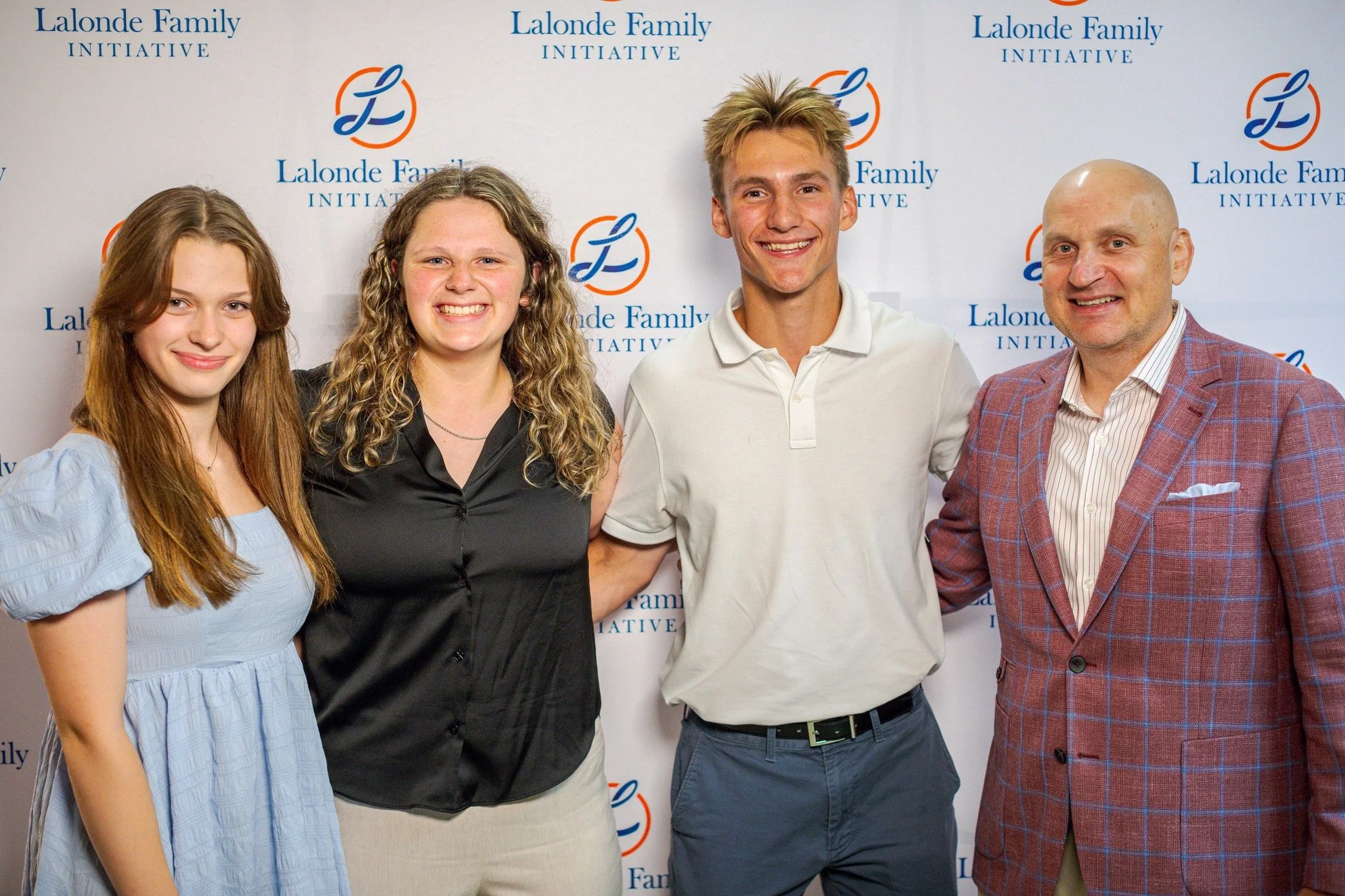 Group photo of four people standing in front of a backdrop with the text 'Lalonde Family Initiative' repeated multiple times and a logo. The group includes two women and two men smiling, dressed in casual and business casual attire.