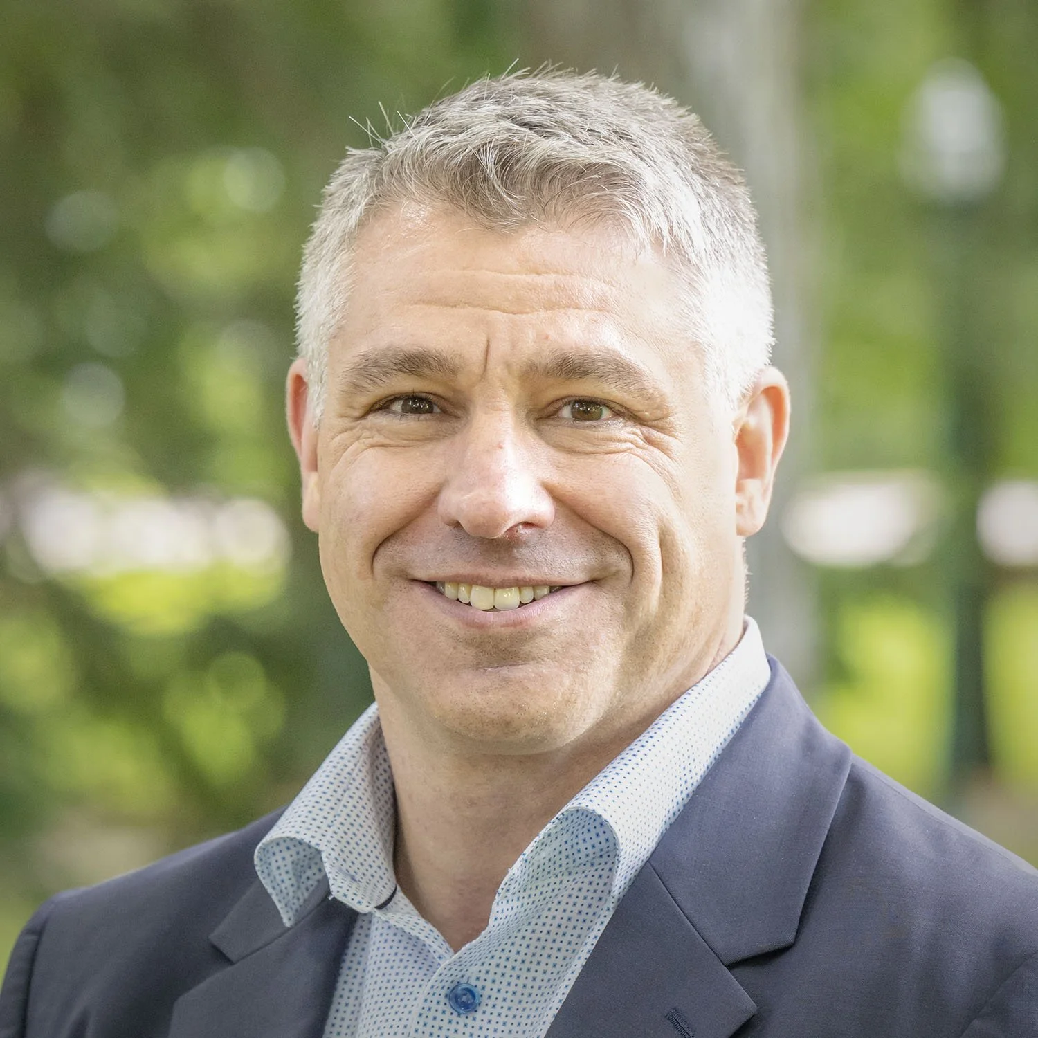 Headshot of a smiling middle-aged man in a suit outdoors with blurred green trees in the background.