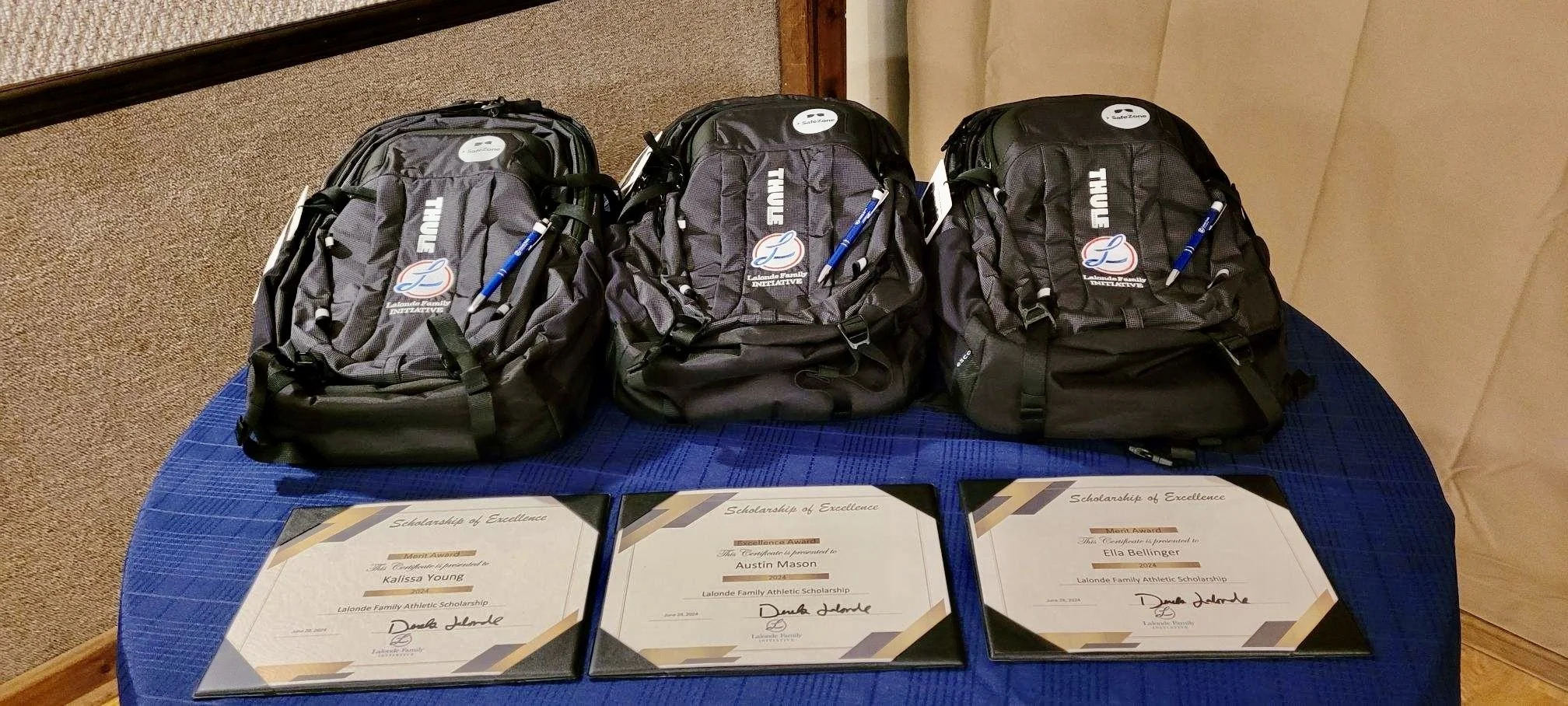 Three black backpacks with 'Thule' logos and blue pens attached, arranged on a blue tablecloth. Below the backpacks, three certificates with signatures and headings that read 'Scholarship of Excellence' and 'Lalonde Family Athletic Scholarship' are displayed.