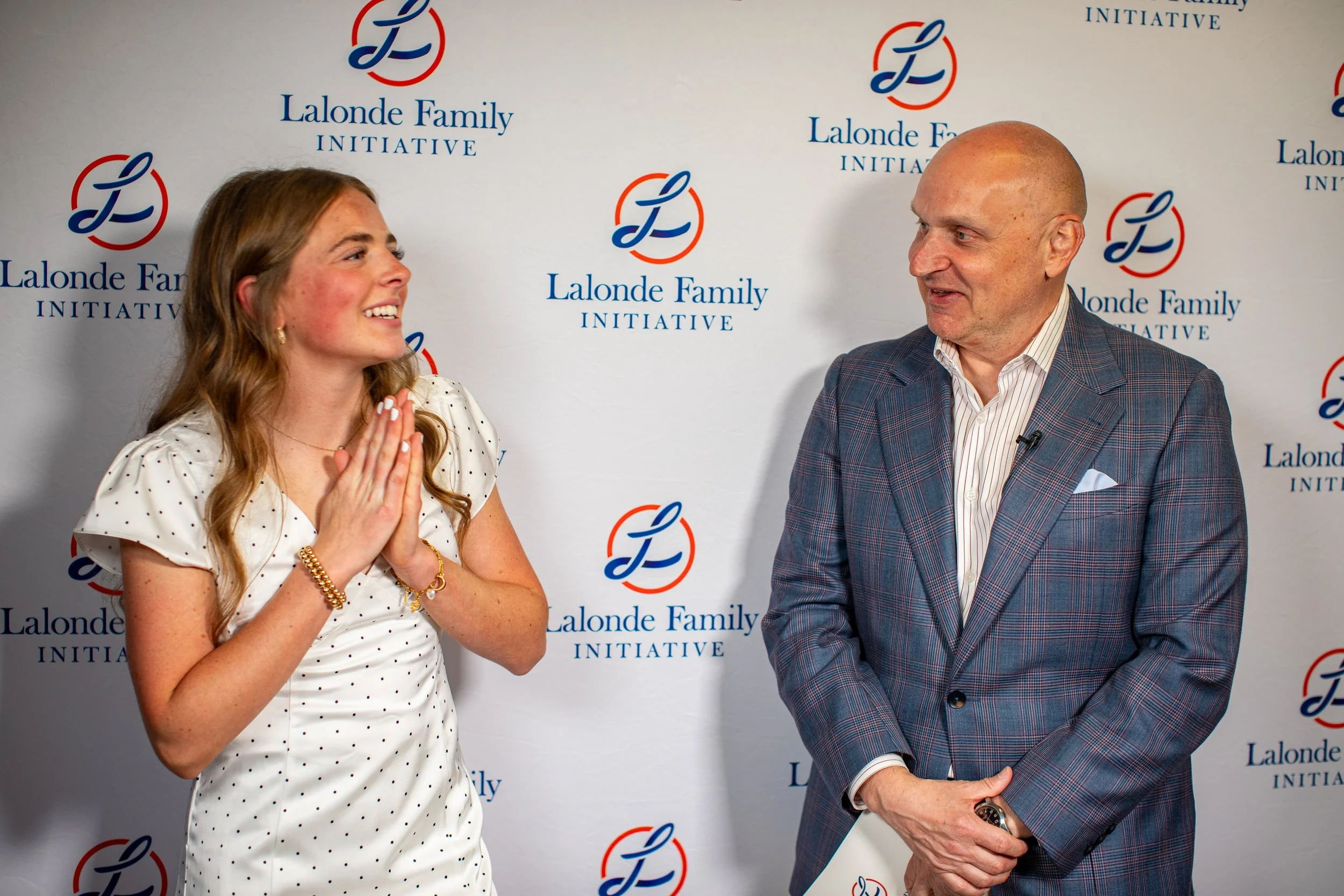 A woman and a man are talking and smiling at each other in front of a backdrop with the logos of Lalonde Family Initiative. The woman has long wavy hair, is wearing a white dress with black polka dots, and gold jewelry. The man is bald, wearing a plaid suit jacket, a white shirt, and holding a phone in his left hand.