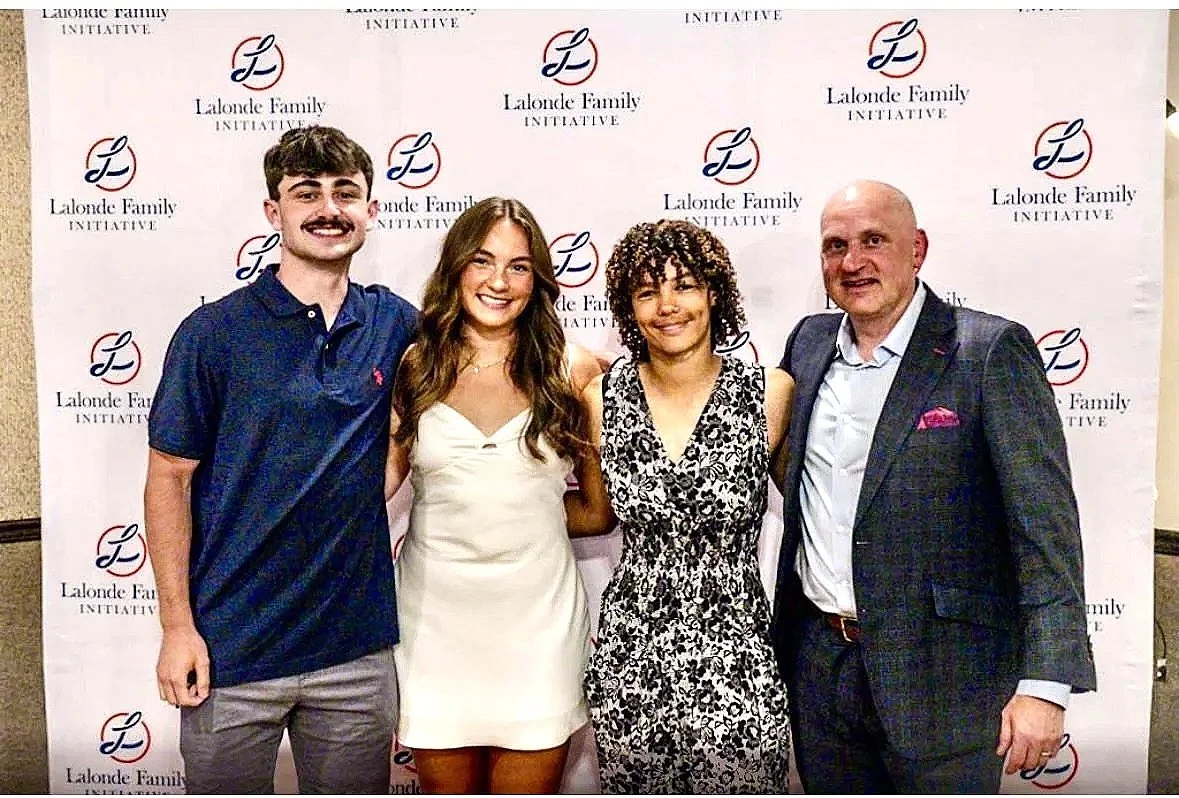 Four people standing together in front of a backdrop with the Lalonde Family Initiative logo. From left to right: a young man in a navy polo shirt and gray pants, a young woman in a white dress, a woman with curly hair in a black and white patterned dress, and a bald man in a dark plaid suit with a white shirt and pink pocket square.