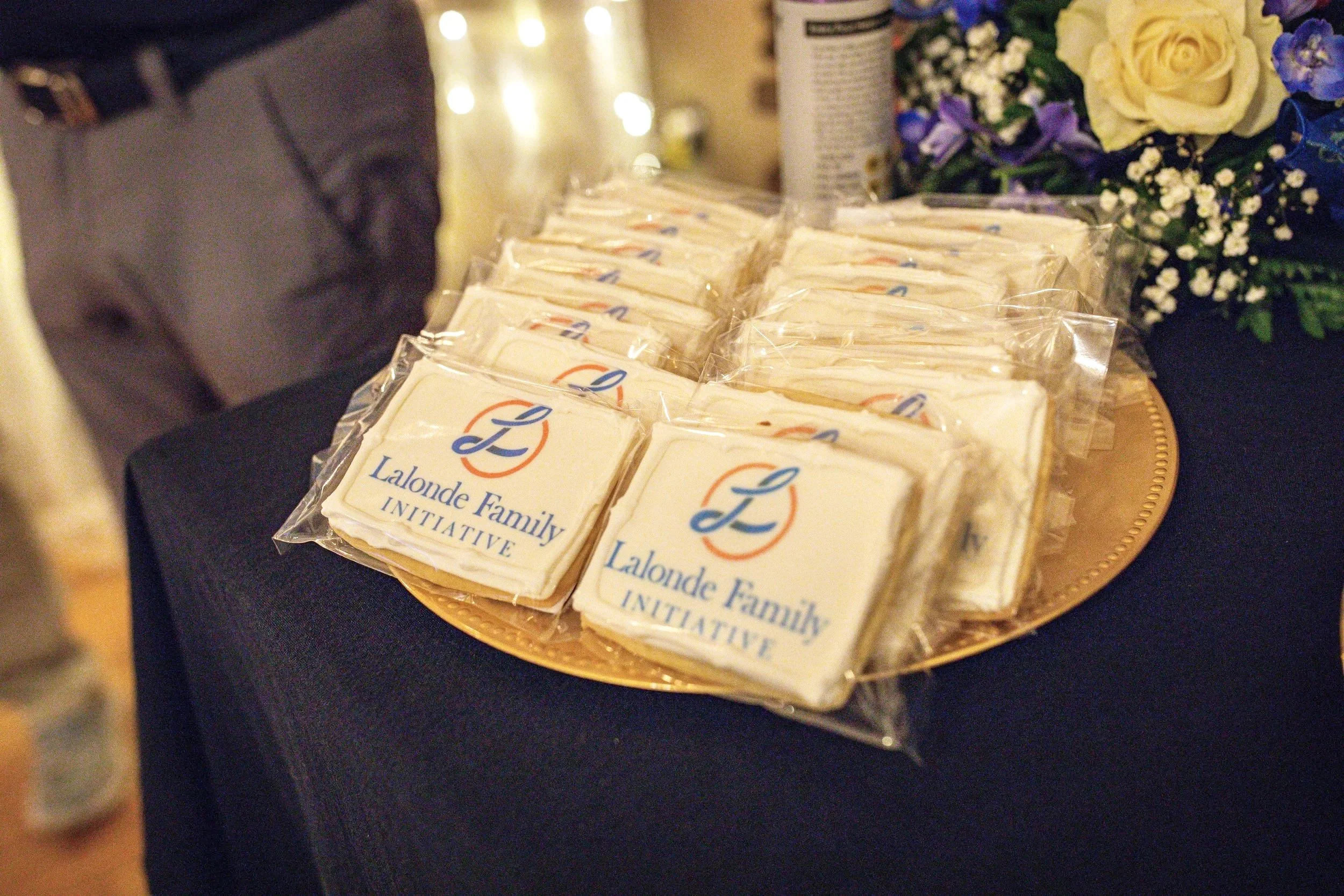 A tray of cookies wrapped in plastic, with a label that reads "Lalonde Family Initiative". In the background, there are flowers and a blurred out table with a can.