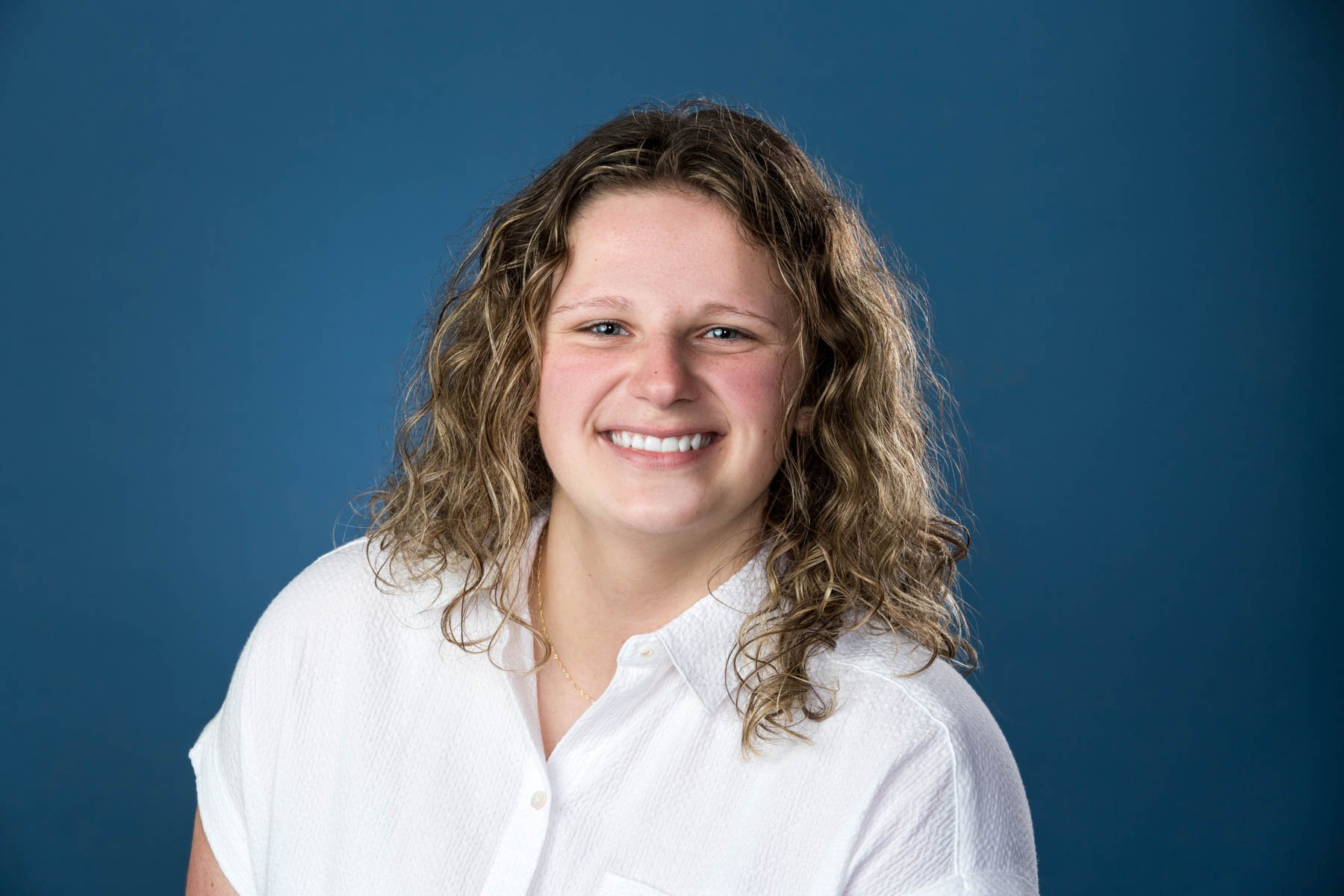 Portrait of a smiling young woman with curly light brown hair, wearing a white button-up shirt against a blue background.