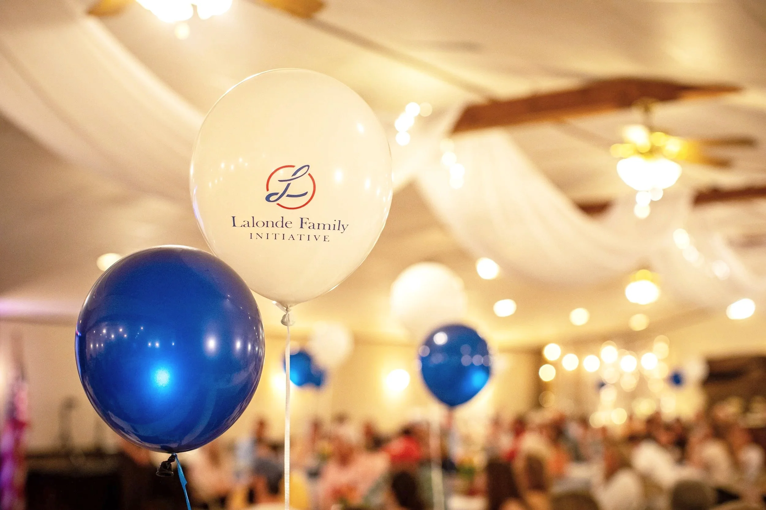 A white balloon with the text 'Lalonde Family Initiative' and a logo, along with blue balloons, hanging from the ceiling during an event with many blurred lights and people in the background.