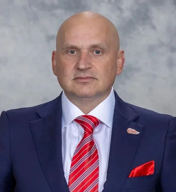 A bald man in a navy blue suit with a white dress shirt and a red and white striped tie, standing against a gray background.