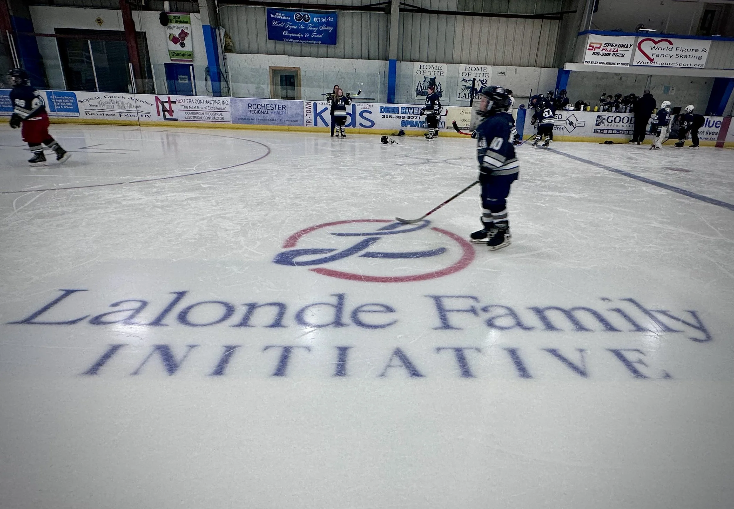 Inside an ice skating rink with players practicing, some fallen helmets on the ice, and an advertisement banner on the boards.