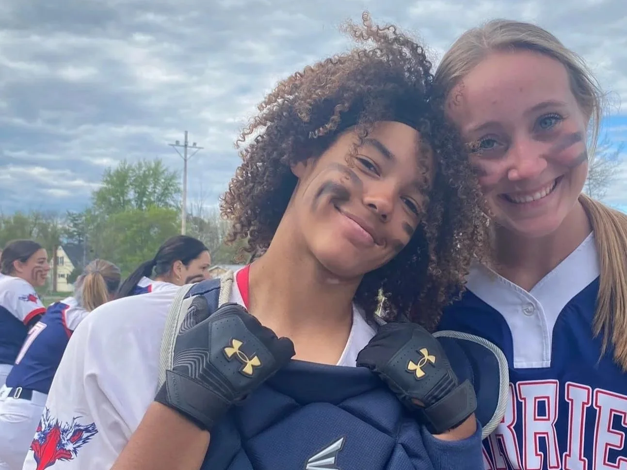 Two female athletes smiling outdoors, one with curly hair and face paint, wearing a football uniform and gloves, the other with straight hair, smiling in a jacket, with a group of people in similar uniforms in the background.