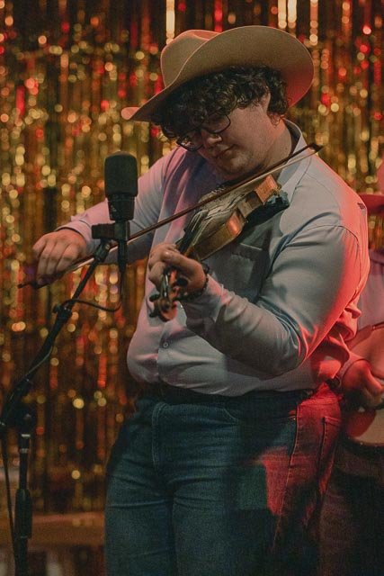 Person playing the violin on stage with a microphone, wearing a large hat and glasses, against a gold glittery backdrop.