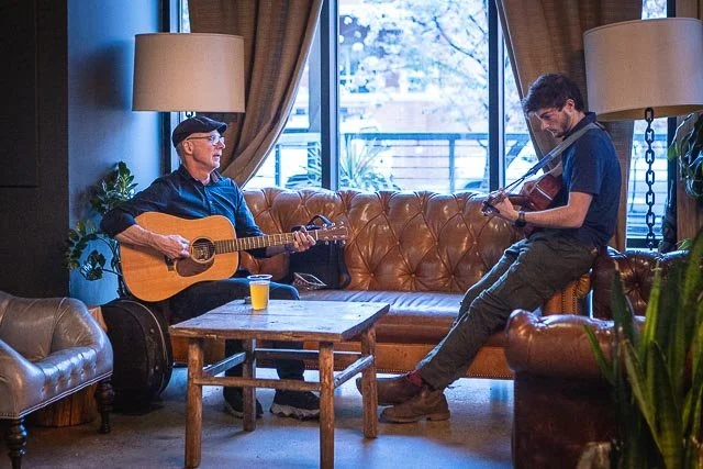 Two men playing acoustic guitars in a cozy living room with large windows, curtains, and plants.