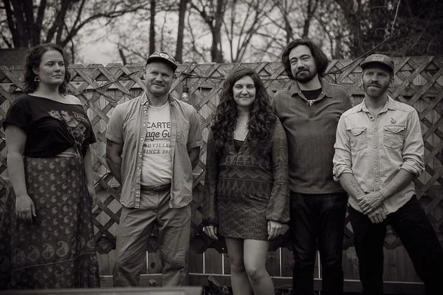 Five people standing outdoors in front of a wooden fence, posing for a photo.