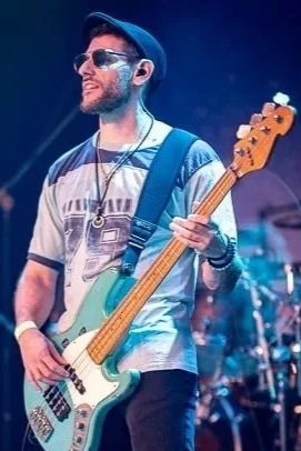 A man wearing sunglasses, a blue cap, and a striped t-shirt playing an electric bass guitar on stage.