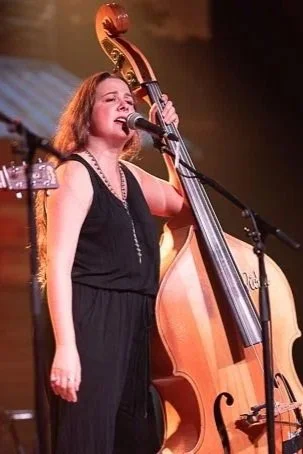 Woman singer performing with a microphone on stage, standing next to a double bass and another guitar.