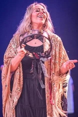 A woman with long blonde hair dancing or performing on stage, holding a tambourine in her right hand, wearing a black dress and a fringed shawl, smiling with her eyes closed.