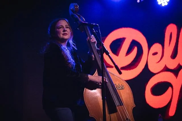 A woman playing an upright bass on stage with a neon red sign in the background reading 'Del'.