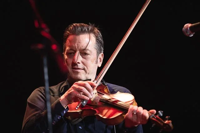 Man playing a violin on stage, with a focused expression, in a dark setting.