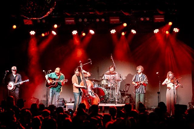 A band performing live on stage with six members playing various instruments under red stage lighting, with an audience in front.