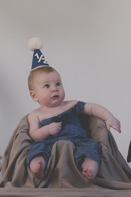 Baby wearing a birthday hat sitting on a chair wrapped in a beige cloth, holding a blue cloth, with a plain light-colored wall in the background.