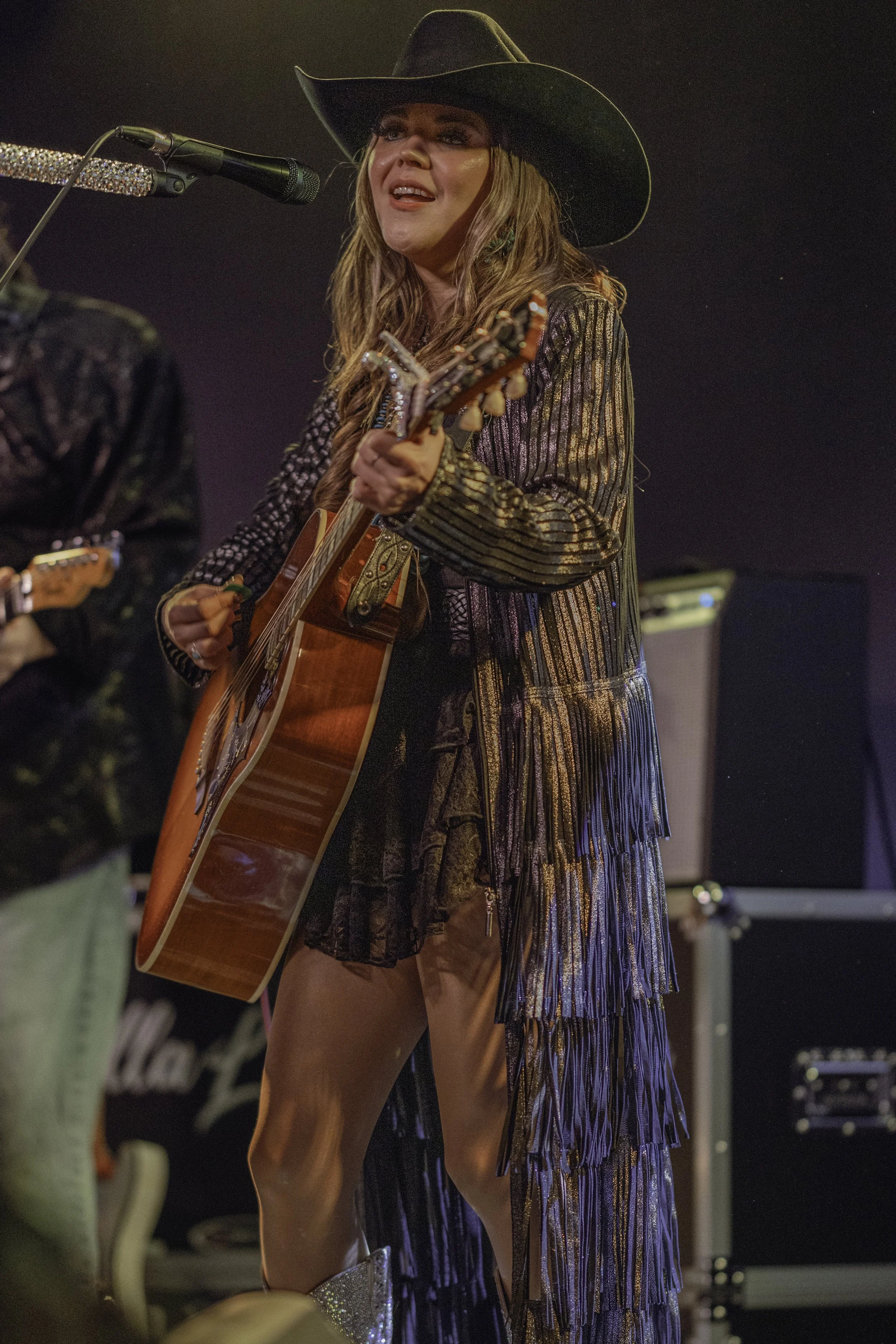 A woman wearing a black cowboy hat and a shiny, metallic dress is playing an acoustic guitar and singing into a microphone at a live music performance.