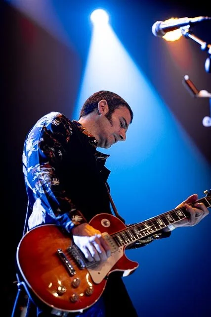Male guitarist playing an electric guitar on stage under blue and white stage lighting.