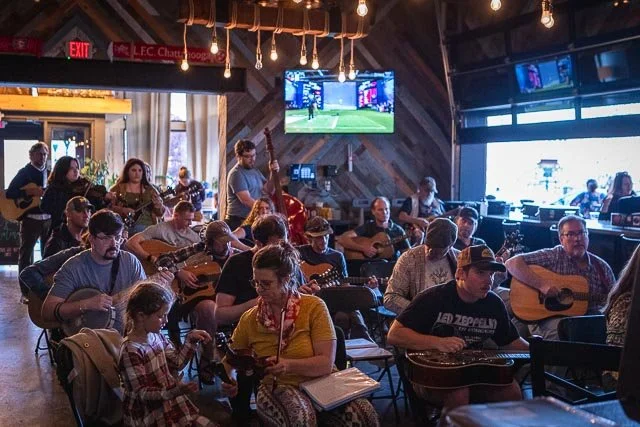 Group of people playing guitars and singing at a restaurant or bar with large windows and TV screens showing sports