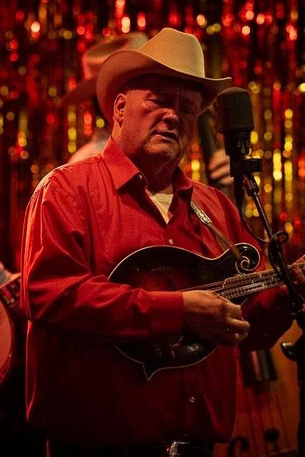 Man in cowboy hat playing mandolin on stage with colorful tinsel backdrop.