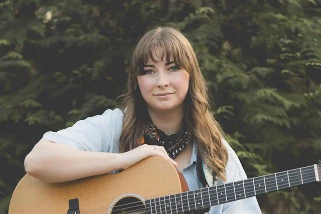 Young woman with long brown hair holding an acoustic guitar outdoors with greenery in the background.