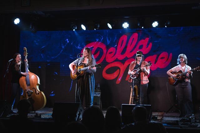 Four women performing on stage with musical instruments, with a large sign that says "Della Mae" behind them, indicating they are performing as the band Della Mae.