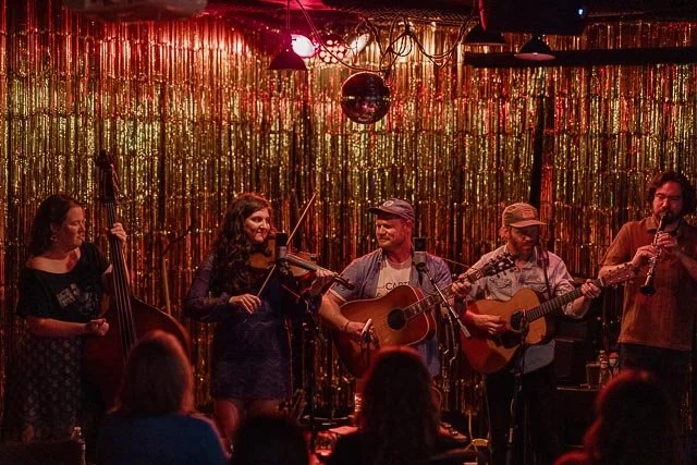 Musicians performing on stage in a small venue with gold glitter backdrop, playing instruments including viola, guitars, and saxophone, with audience members in the foreground.