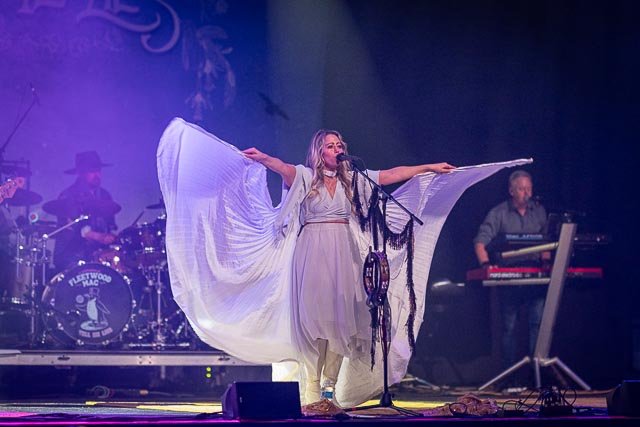 A female singer with large flowing white sleeves performs on stage with a band, including a drummer and keyboardist, at a concert or music venue.