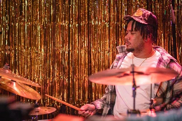 A man playing drums in front of a gold tinsel curtain, wearing a plaid shirt and a cap.