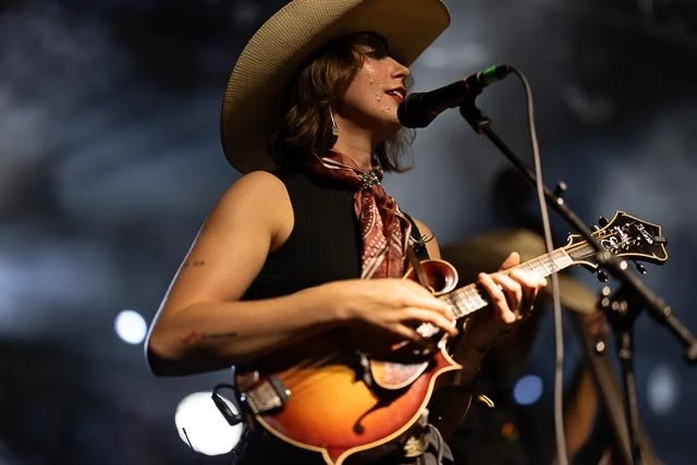 A woman wearing a large cowboy hat and bandana playing a small guitar or ukulele and singing into a microphone on stage.