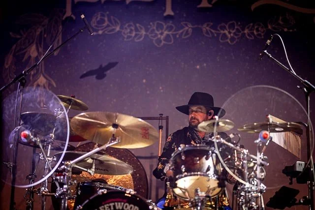 A man playing a drum set on stage with microphones, stage lights, and a backdrop decorated with flowers and a bird silhouette.