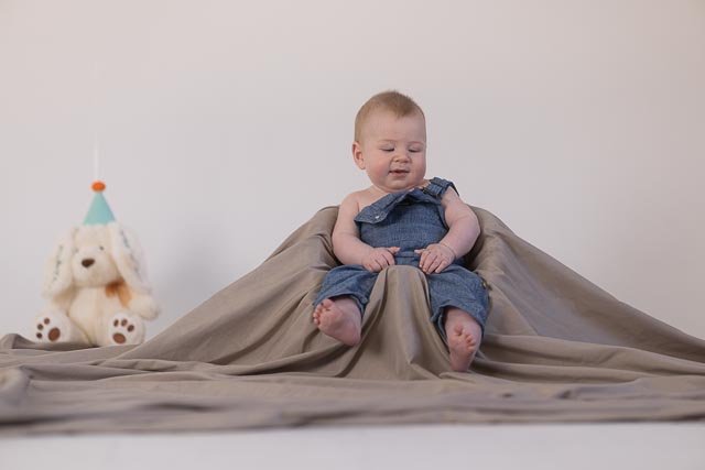 A baby sitting on a beige fabric backdrop with a white stuffed bear wearing a blue party hat in the background.