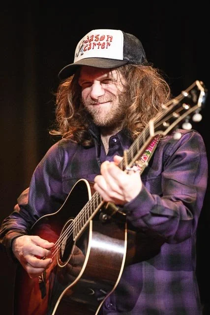 A man with long hair and a beard playing an acoustic guitar, wearing a purple plaid shirt and a white trucker hat with red text that reads 'Jason Isbell' on the front.