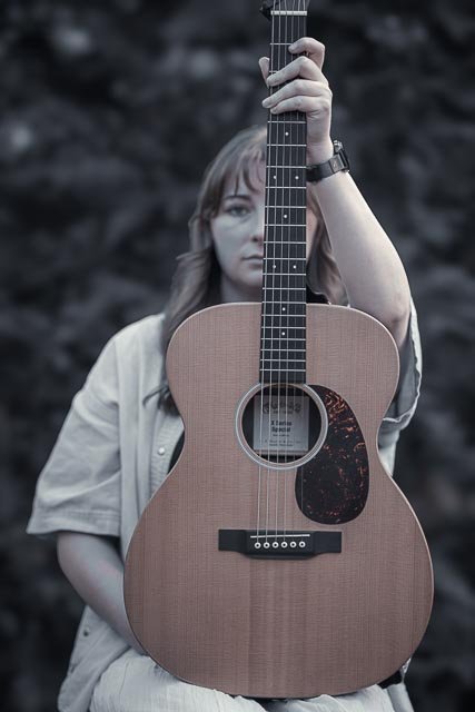 A woman holding an acoustic guitar vertically in front of her face, with only her eyes and part of her hair visible, outdoors with blurred rocks in the background.