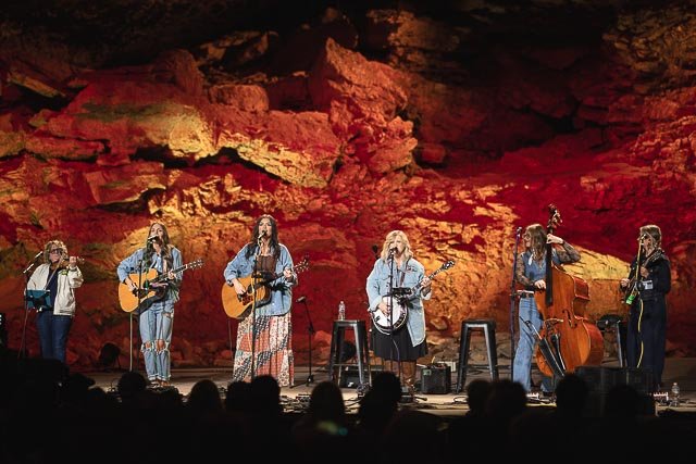 A musical band performing on stage with a rocky, illuminated background. The band includes six women playing instruments such as guitars, banjo, upright bass, and singing.