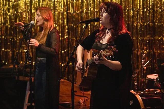 Two women singing and playing guitar on stage with gold sequin backdrop.