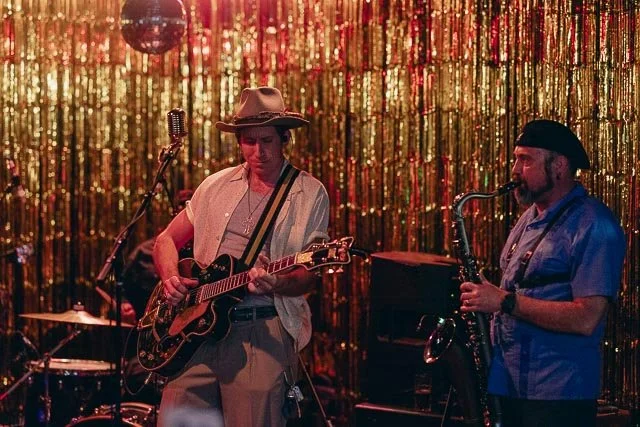 Two musicians performing on stage with a gold glittering backdrop. One plays guitar and wears a hat, while the other plays saxophone.
