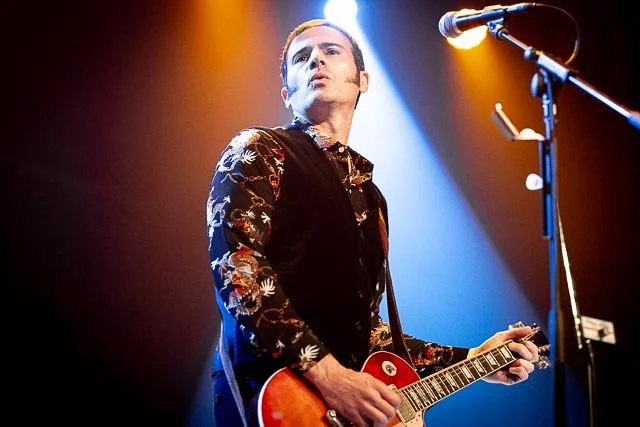 Male musician playing an electric guitar on stage with colorful lighting.