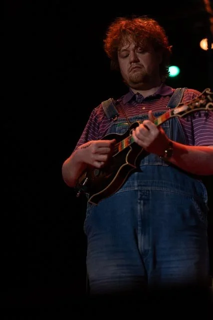 A person with curly hair and a beard playing a guitar on stage, wearing overalls and a striped shirt.