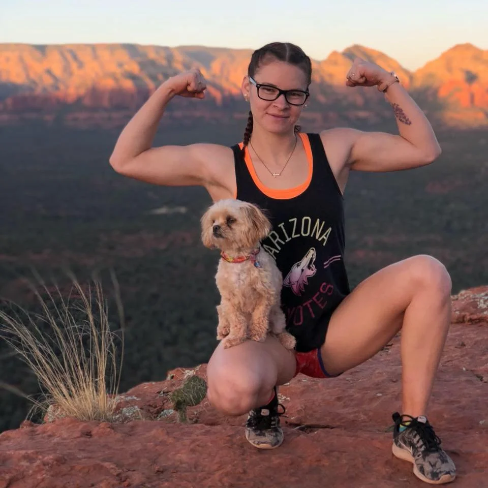 A woman in athletic gear flexing her biceps on a rocky ledge with mountains in the background, holding a small dog.