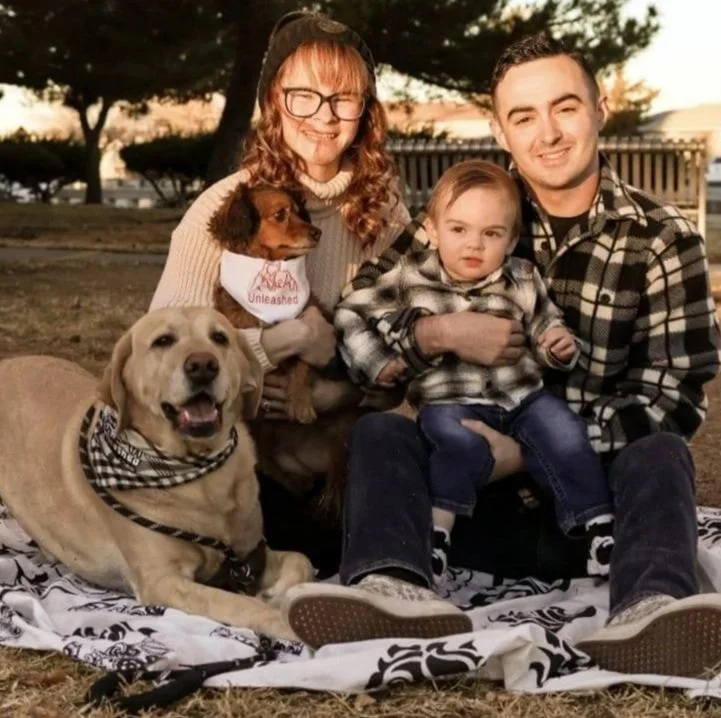 A family of three with two dogs sitting on a blanket outdoors during sunset. The mother has glasses and curly hair, holding a small brown dog. The father has short dark hair, and is holding a young child with blonde hair. The larger dog, a yellow Labrador, is lying on the blanket and wearing a bandana.