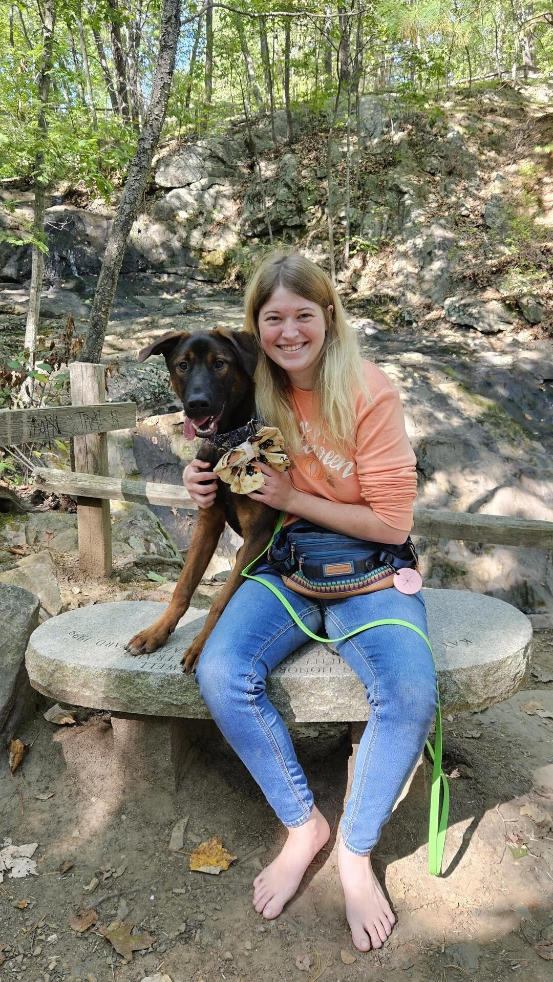 A young woman with long blonde hair, smiling, sitting barefoot on a stone bench in a wooded area, holding a black and brown dog with a bandana around its neck.