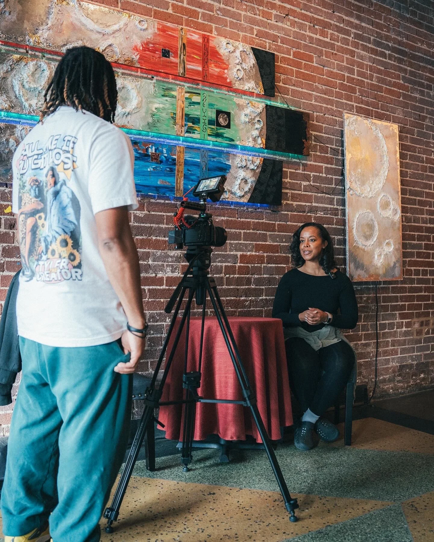 A woman is sitting at a table with a red tablecloth, being recorded by a person with a camera on a tripod. The scene is set against a brick wall with abstract artwork hanging on it.