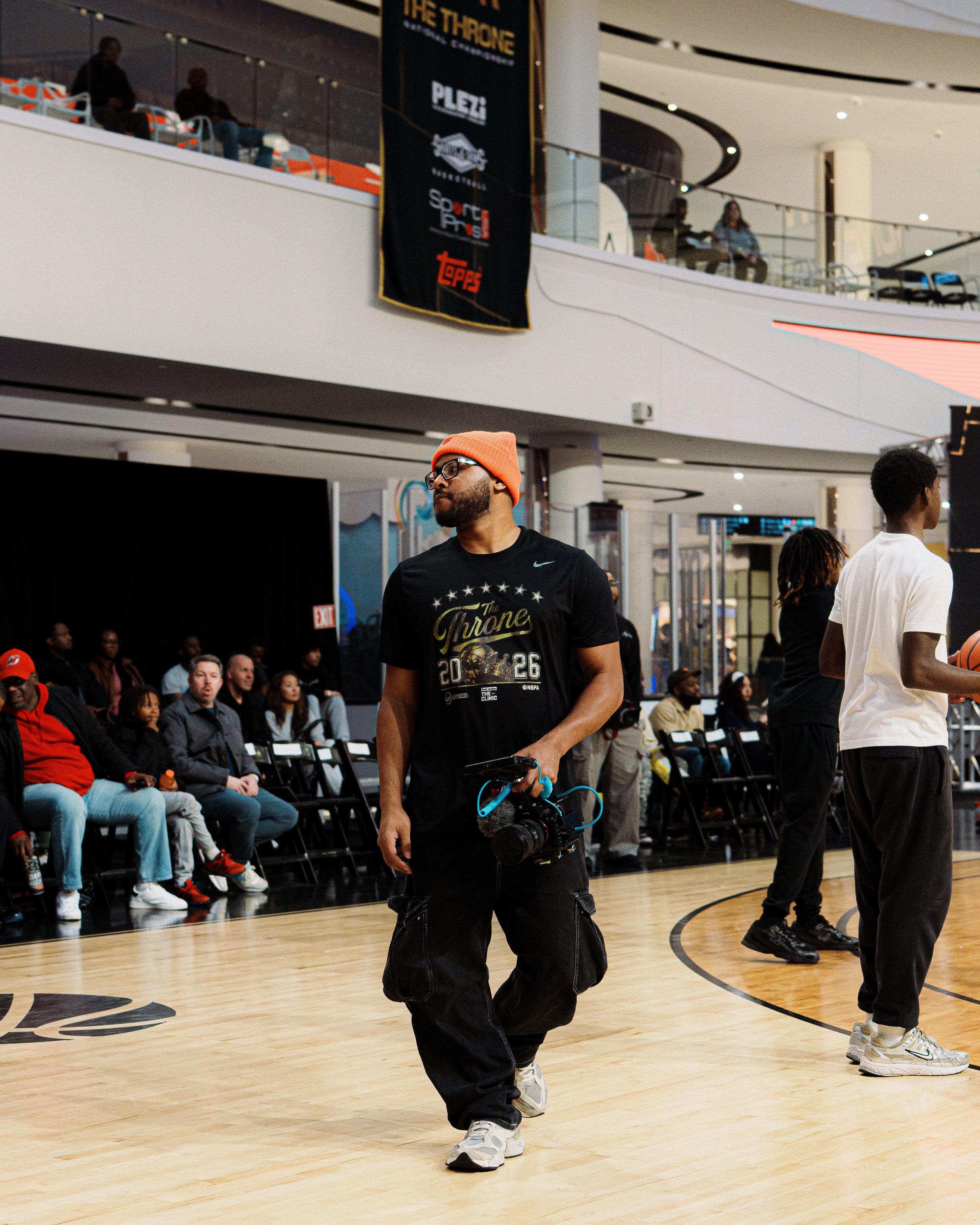 Man in black Nike shirt, orange beanie, glasses, and white sneakers walking on basketball court, holding camera, with audience seated in background and banners above.