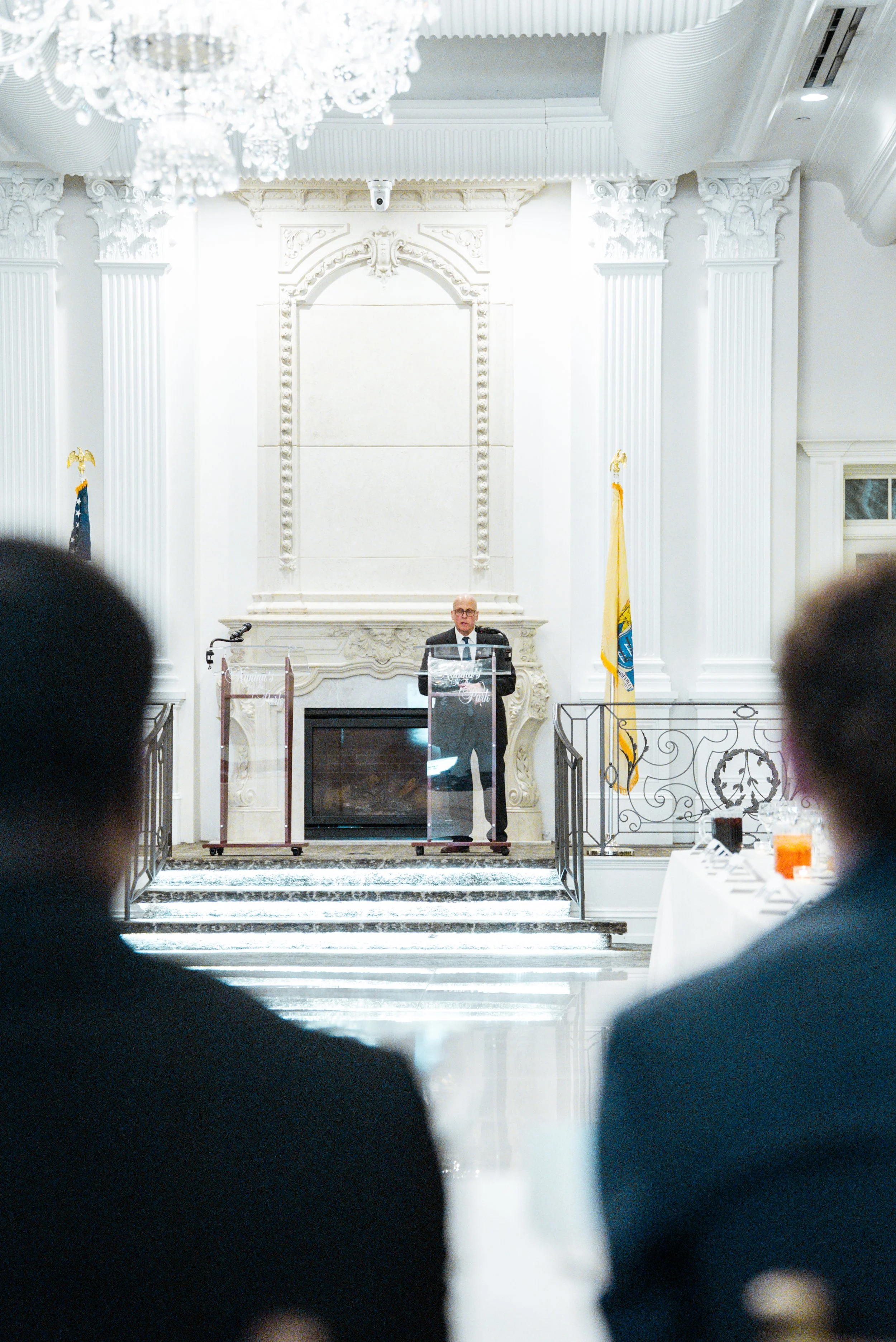 A man in a suit speaking at a podium with a screen and flags in the background, in a white decorated hall with columns and a chandelier.