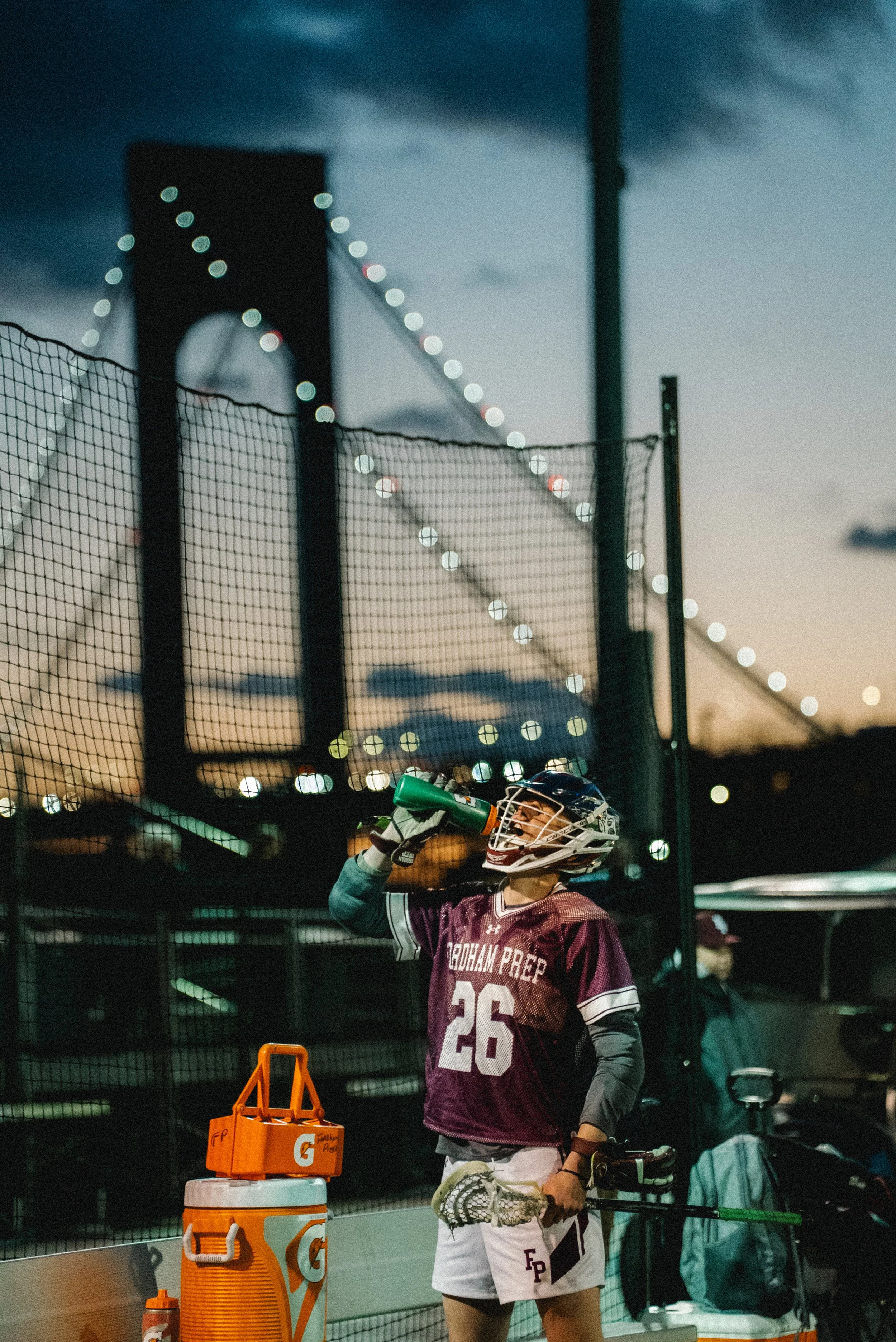 A male lacrosse player wearing a maroon and white uniform with 'FGUAM PREP' and the number 26 is drinking from a green Gatorade bottle during a game at dusk, standing near a bench with hydration supplies, behind a sports fence, with a bridge and darkening sky in the background.